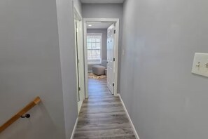 A narrow hallway with a wooden handrail, a light switch, and a door leading to a room with a window and a round object on the floor.