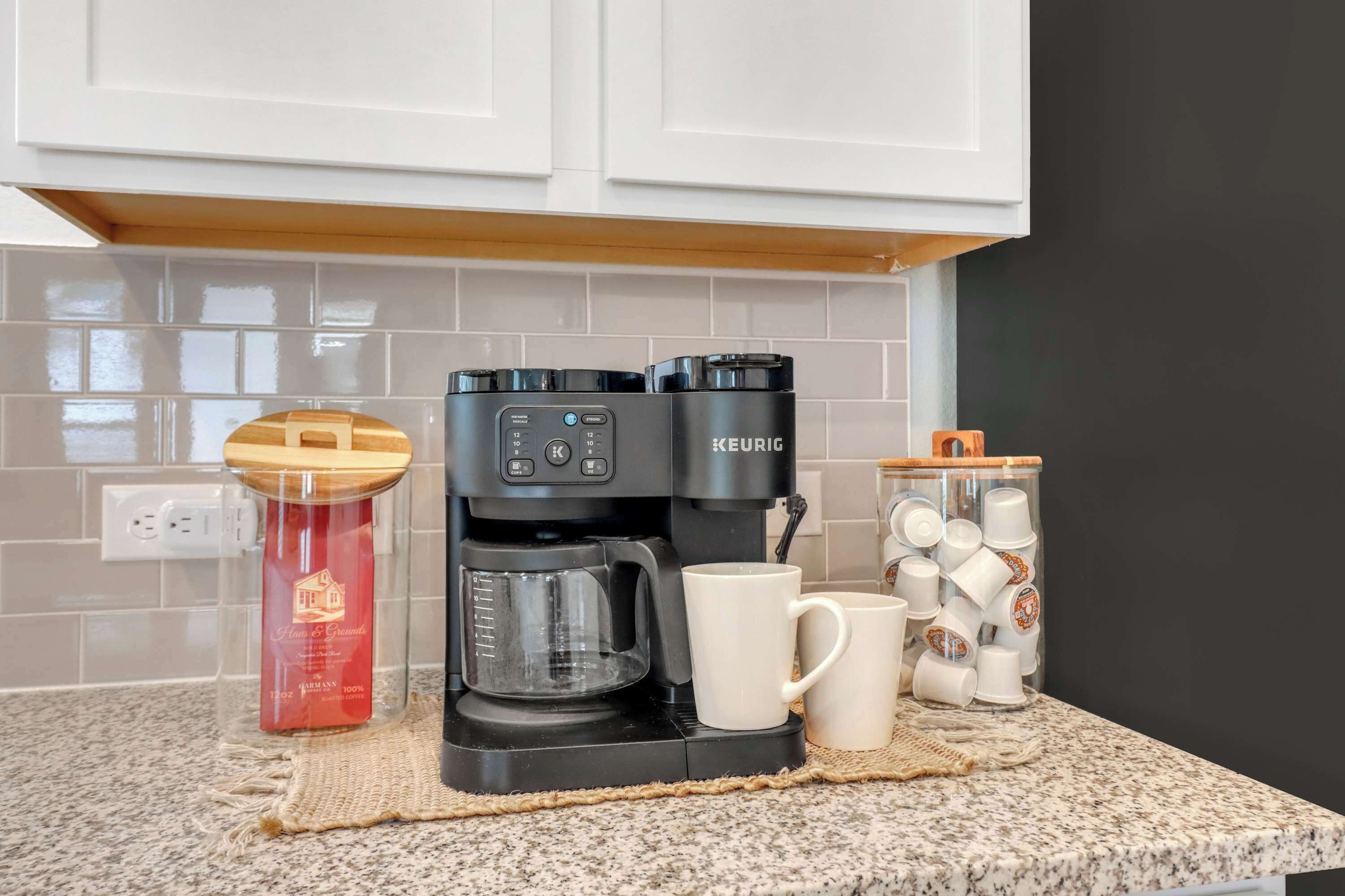 A kitchen with a Keurig coffee maker, a glass jar with a red label, and a container of white mugs.