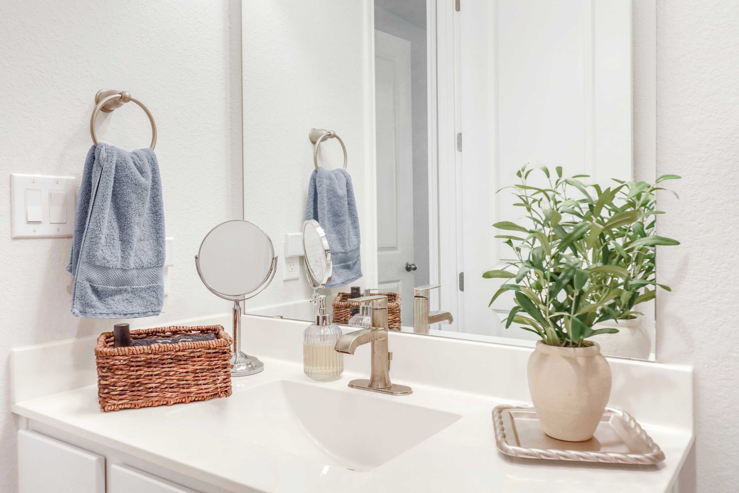 Une salle de bain avec un lavabo blanc, un miroir, deux serviettes, un panier en osier, un distributeur de savon et une plante en pot.