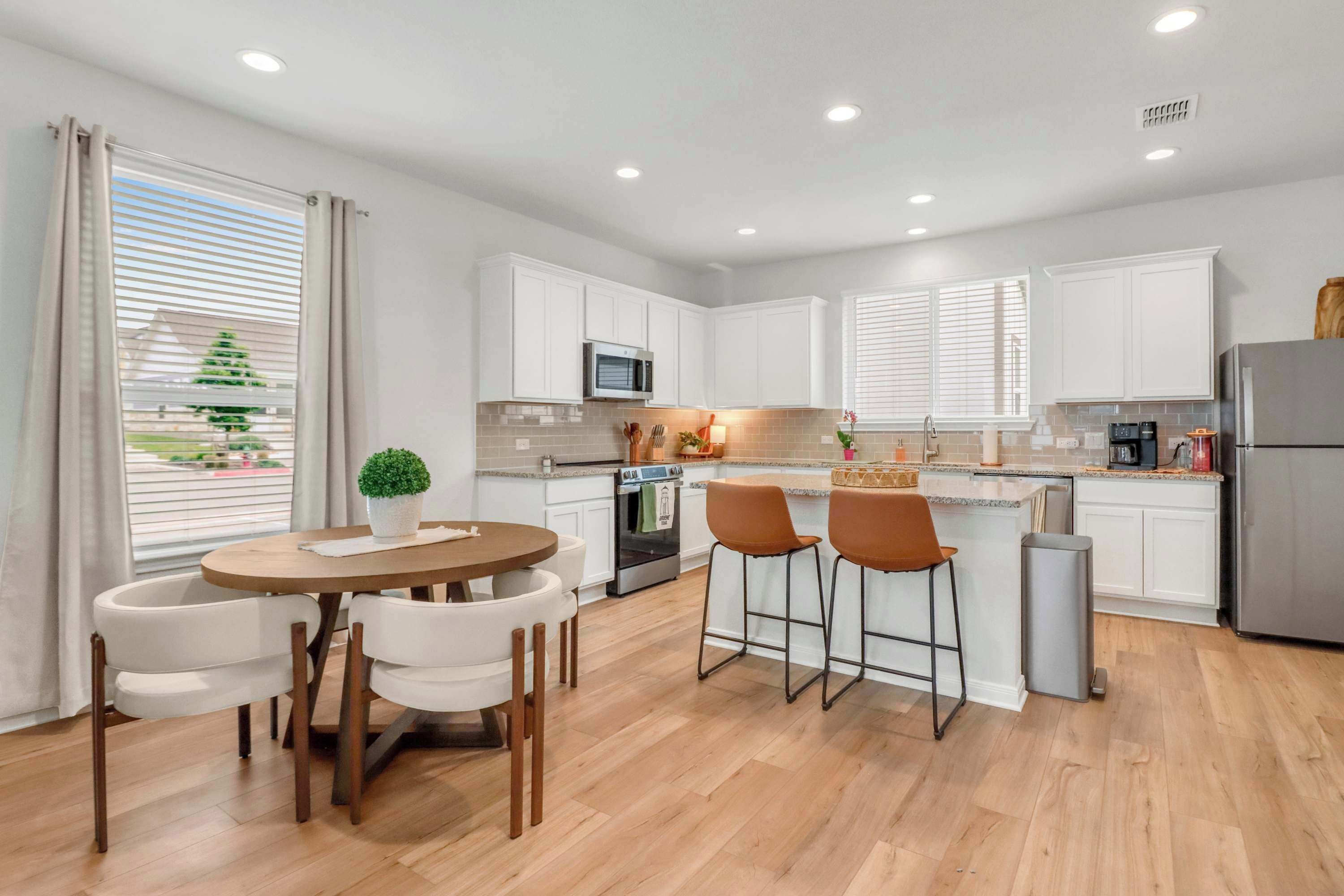 Una cocina moderna con armarios blancos, una isla central con taburetes de bar, una mesa de comedor con sillas y vistas al exterior a través de amplios ventanales.