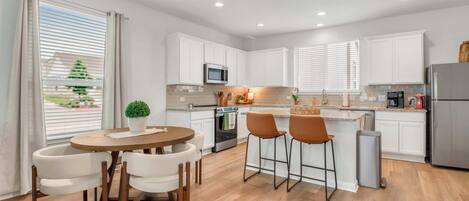 Una cocina moderna con armarios blancos, una isla central con taburetes de bar, una mesa de comedor con sillas y vistas al exterior a través de amplios ventanales.