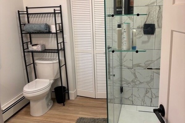 Un baño con inodoro blanco, estante negro, cabina de ducha de vidrio y pared de ducha con mosaicos de mármol.