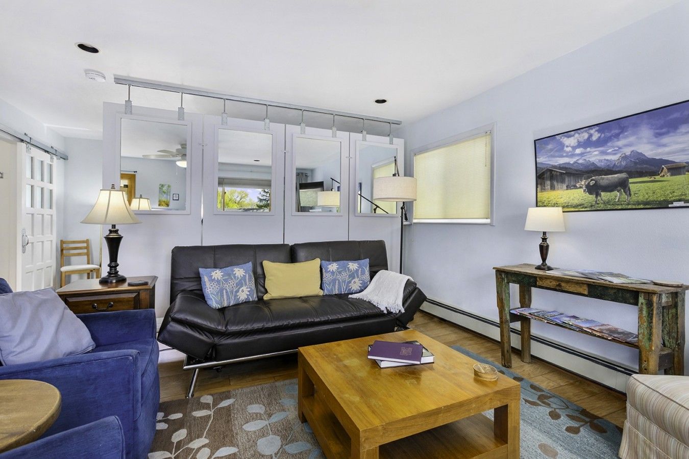 A living room with a black sofa, a wooden coffee table, and a blue armchair. There is a painting on the wall and a lamp on a side table.