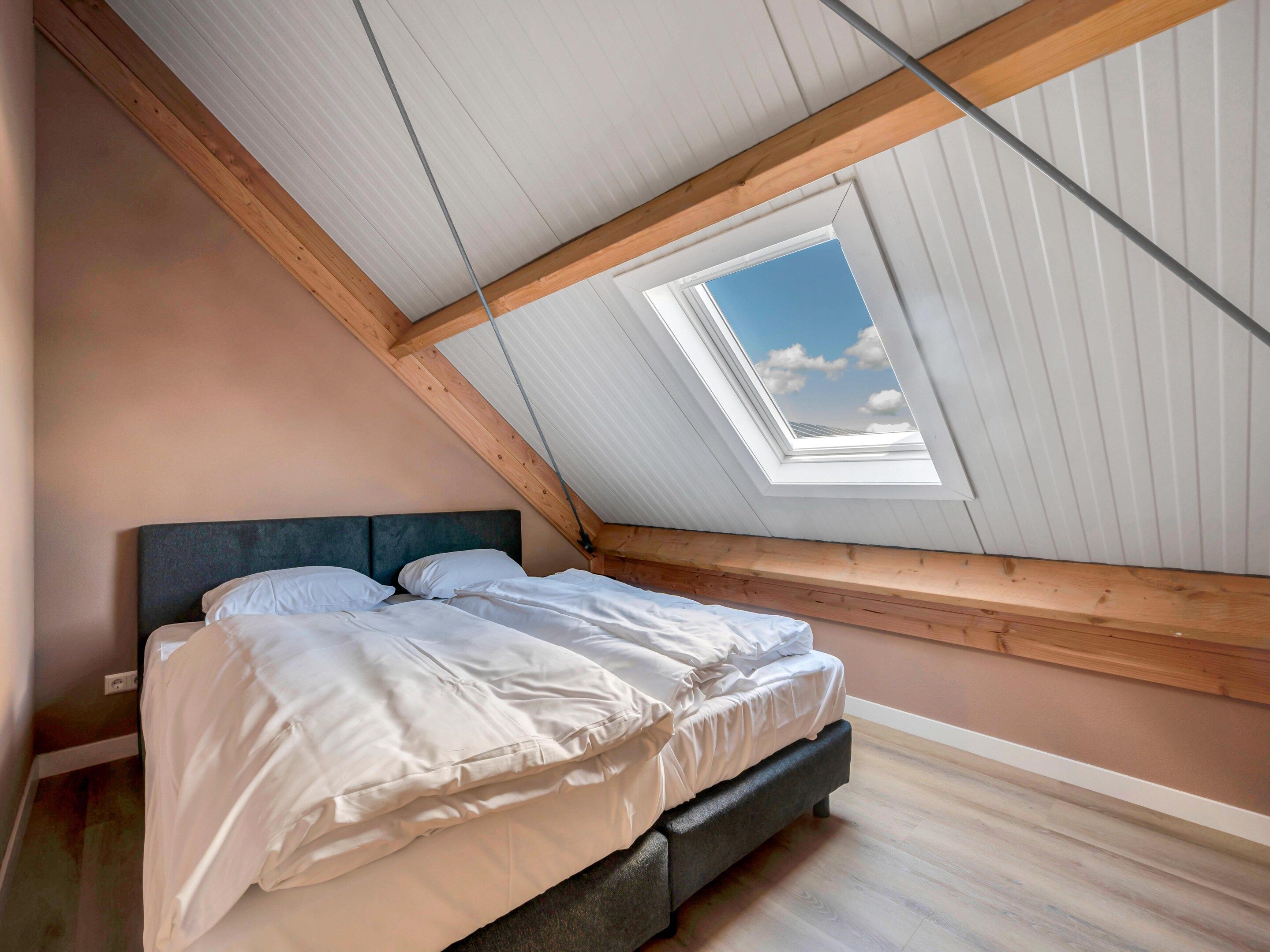 Une chambre avec un plafond en pente, un puits de lumière, un lit avec une literie blanche et des poutres en bois.