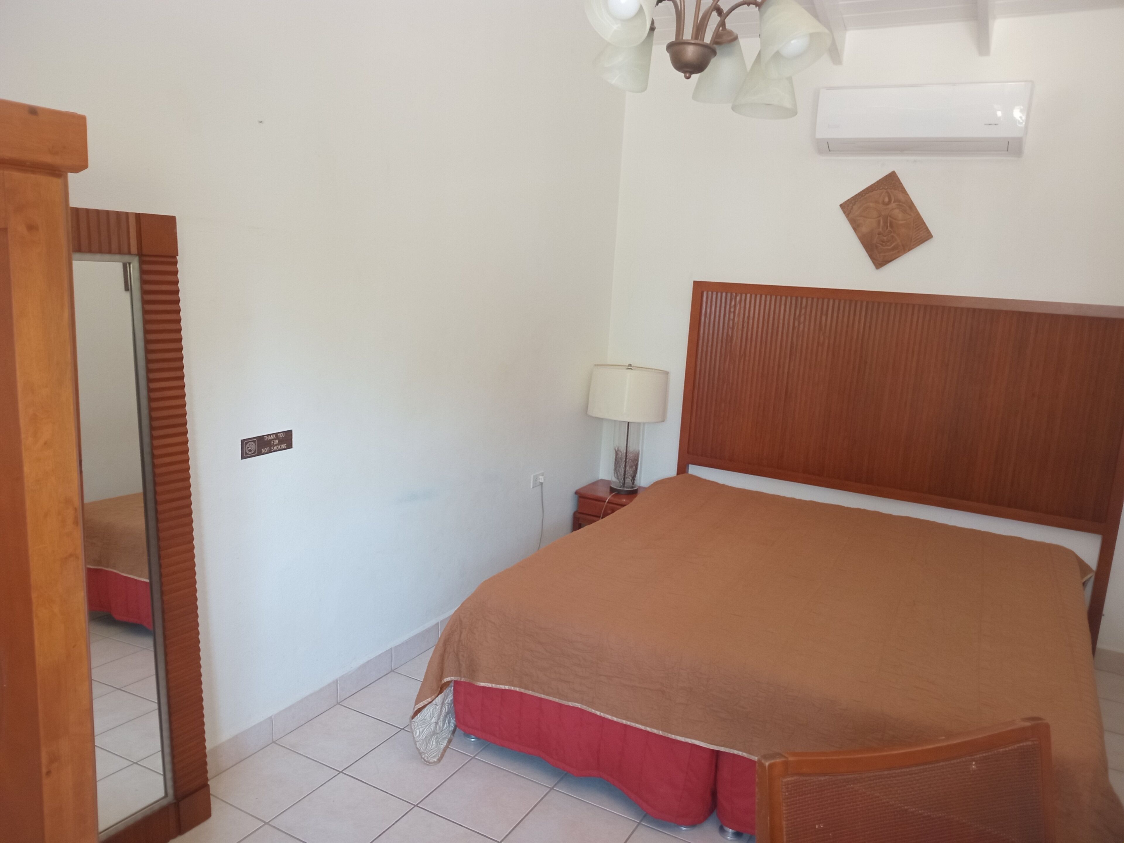 A bedroom with a wooden headboard, a bed with a brown cover, a bedside table with a lamp, a wall-mounted air conditioner, and a mirror.