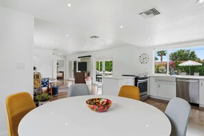 A modern kitchen with white cabinets, a dining table with chairs, and a view of the outdoors. - Mountain View Retreat - Pool, Palms, and Peace (Palm Springs)