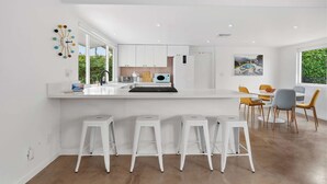 A modern kitchen with white cabinetry, a central island with stools, and a dining area with chairs. - Mountain View Retreat - Pool, Palms, and Peace (Palm Springs)