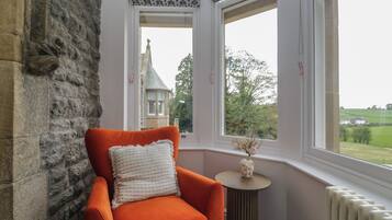 A stone wall, a window with a view of a building and greenery, and an orange chair with a white cushion.