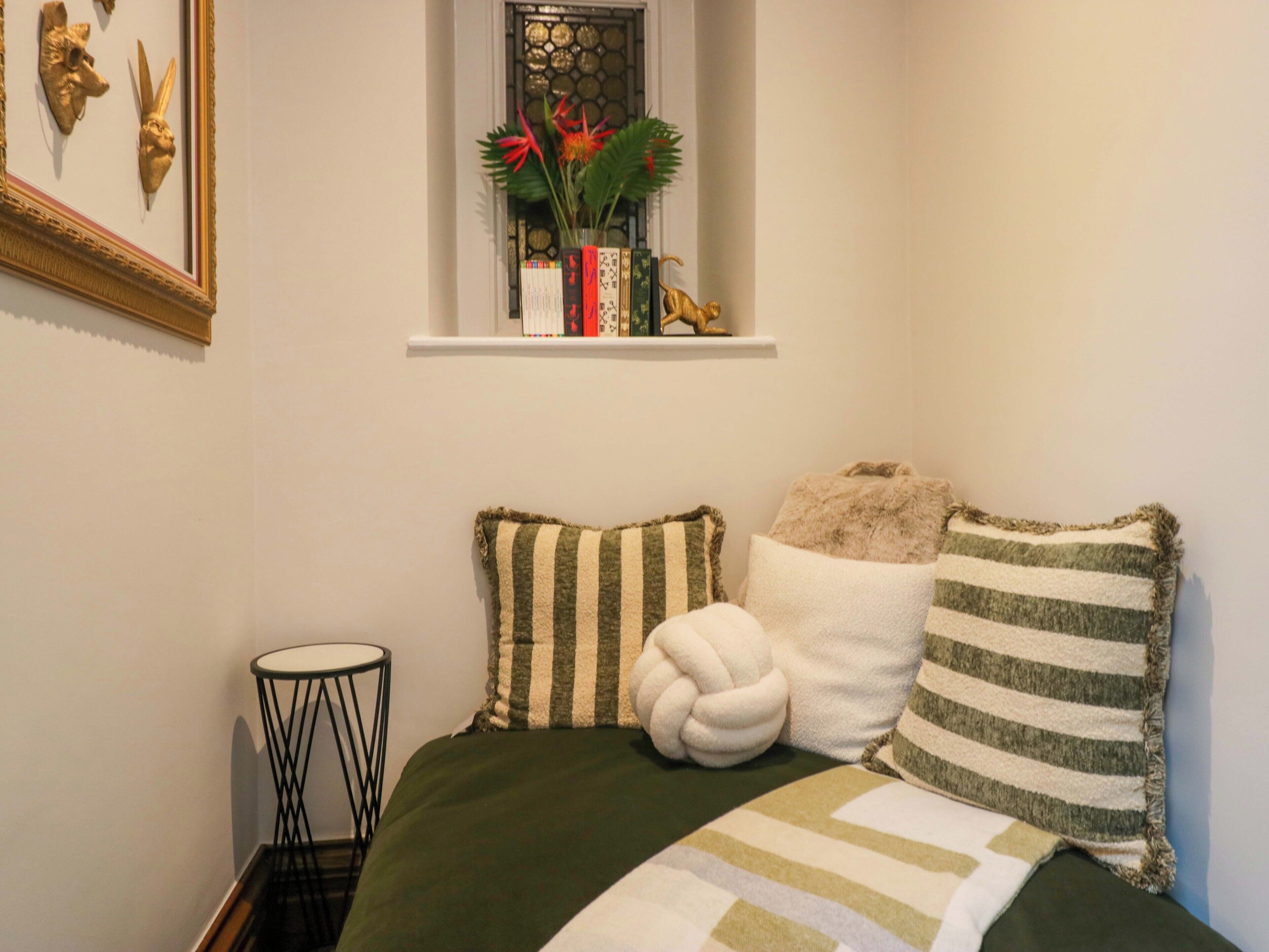 A bedroom with a bed, pillows, a small side table, a window with a plant, and a wall-mounted shelf with decorative items.