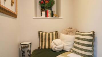 A bedroom with a bed, pillows, a small side table, a window with a plant, and a wall-mounted shelf with decorative items.