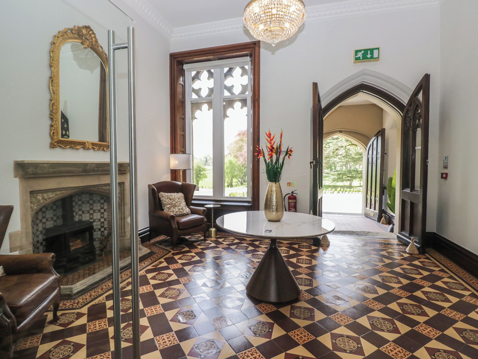 Un hall d’entrée avec un sol à motifs, une cheminée, une table ronde ornée d’un vase, une chaise et un lustre.