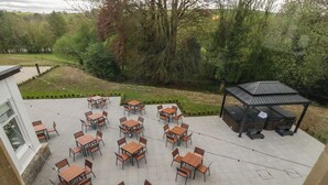 Un patio exterior con varias mesas y sillas de madera, una zona de barbacoa y vistas a un jardín ajardinado.