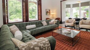 Una sala de estar con un sofá verde, una mesa de centro de vidrio y ventanas grandes con cortinas estampadas.