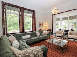A living room with a green sofa, a glass coffee table, and large windows with patterned curtains.