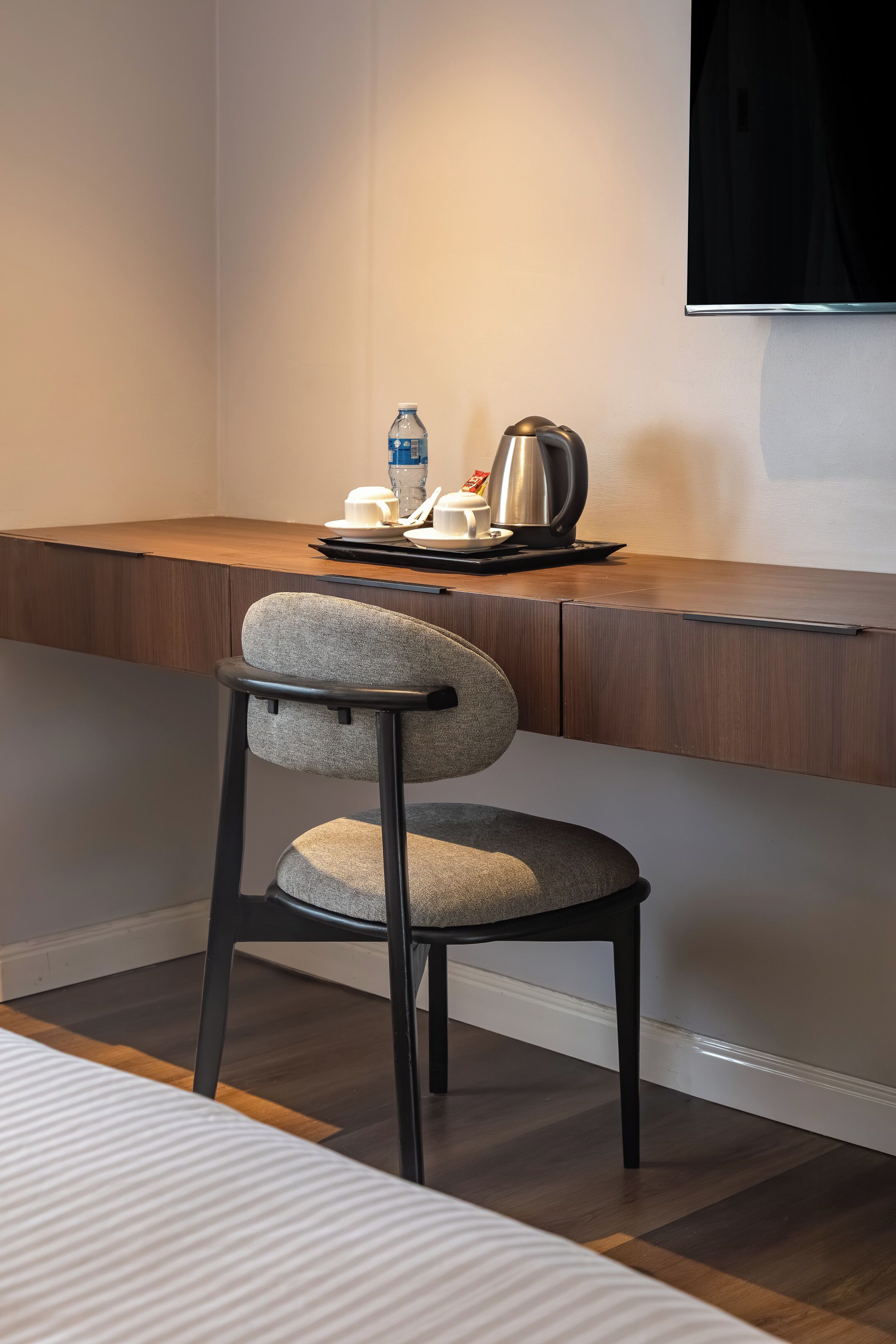 A modern hotel room with a wooden desk, a grey upholstered chair, a flat-screen TV mounted on the wall, and a small table with a kettle, cup, and water bottle.
