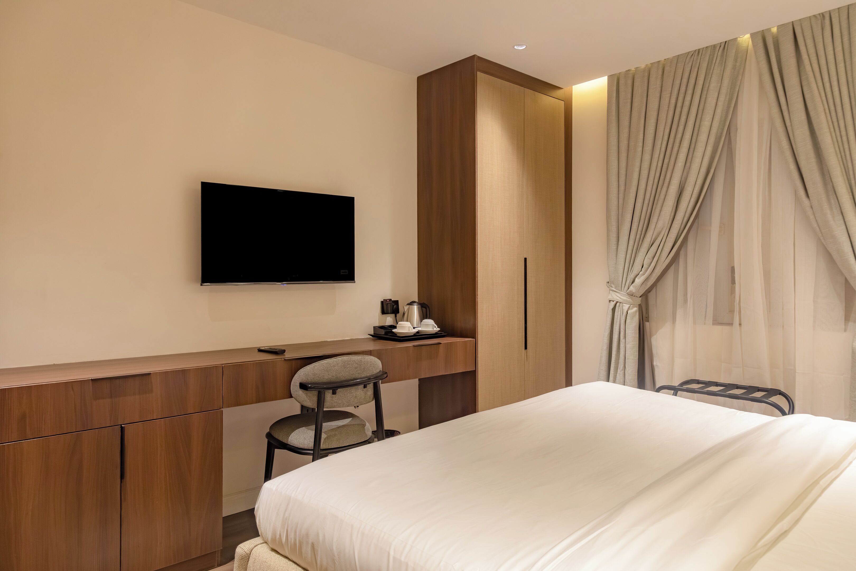 A hotel room with a bed, a desk with a chair, a television mounted on the wall, and a wardrobe.