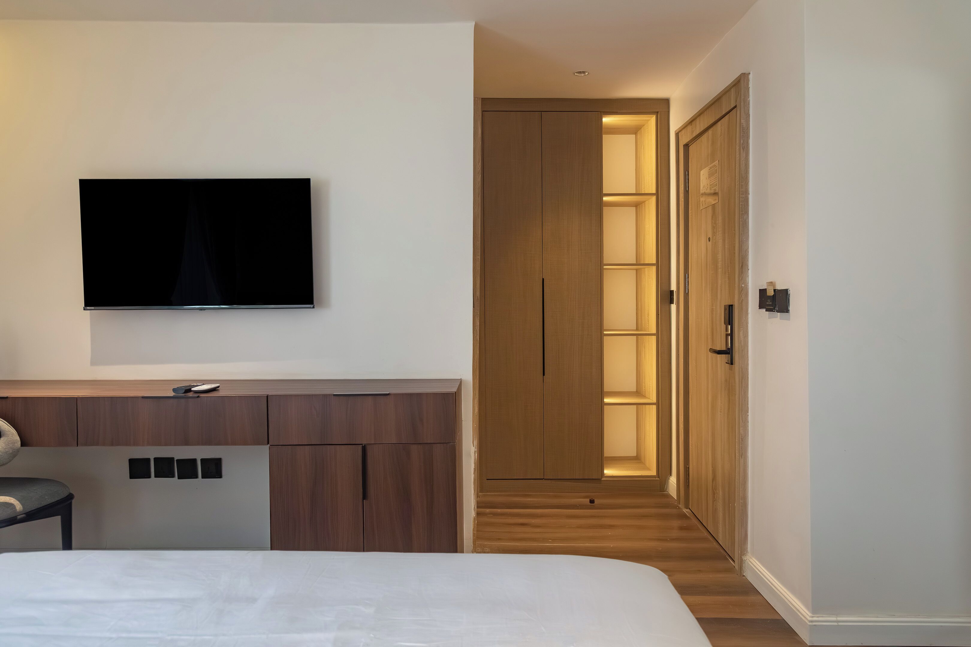 A modern hotel room with a bed, a TV mounted on the wall, a wooden wardrobe, and a hallway leading to a door.