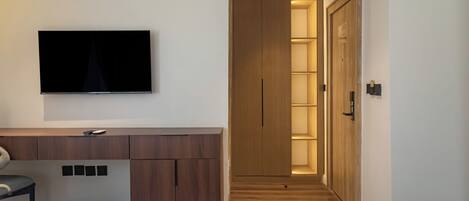 Una habitación de hotel moderna con una cama, un televisor fijado a la pared, un armario de madera y un pasillo que lleva a una puerta.