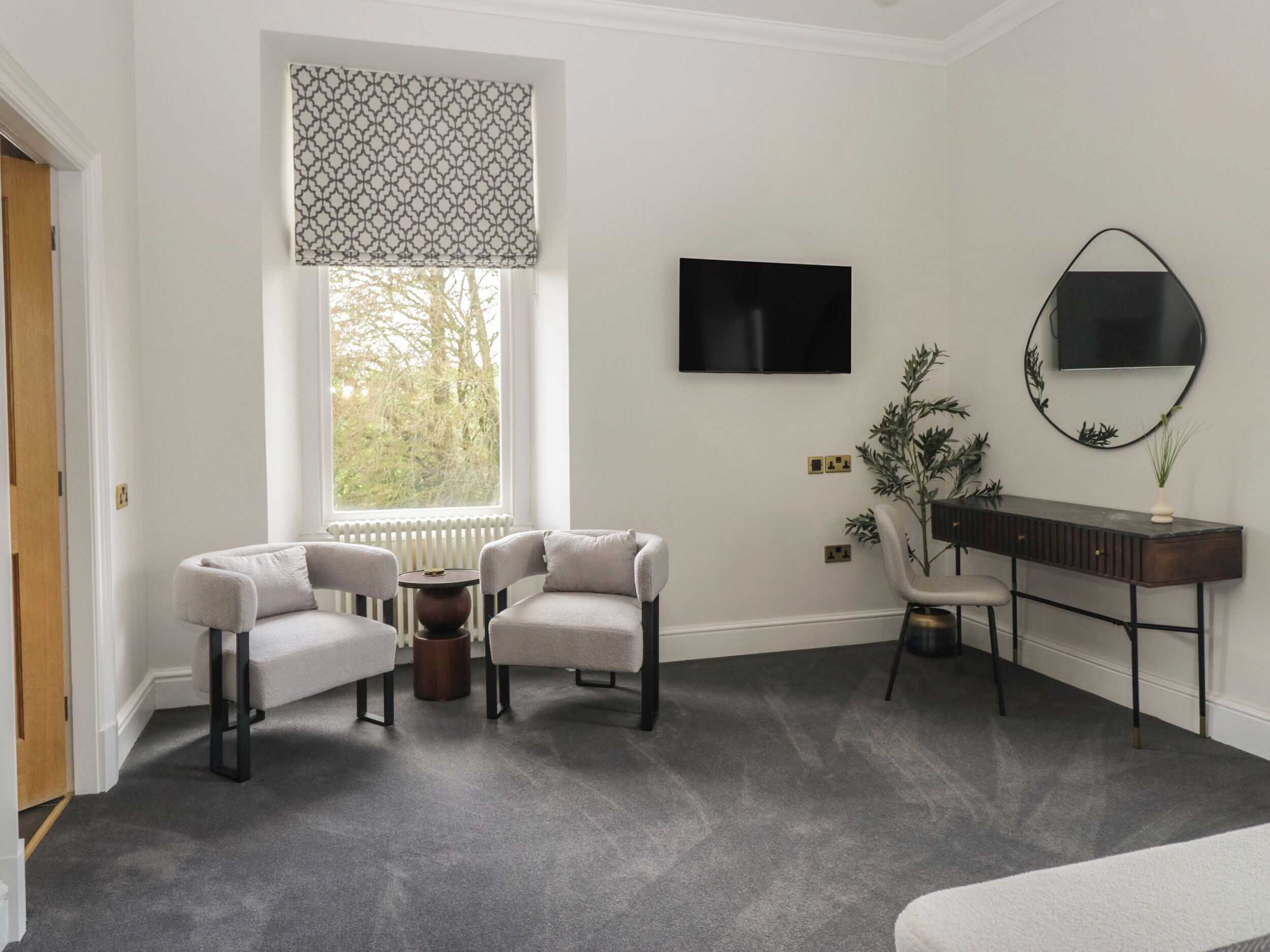 Una habitación con alfombra gris, dos sillones, una mesa redonda, un televisor, un espejo y un radiador.