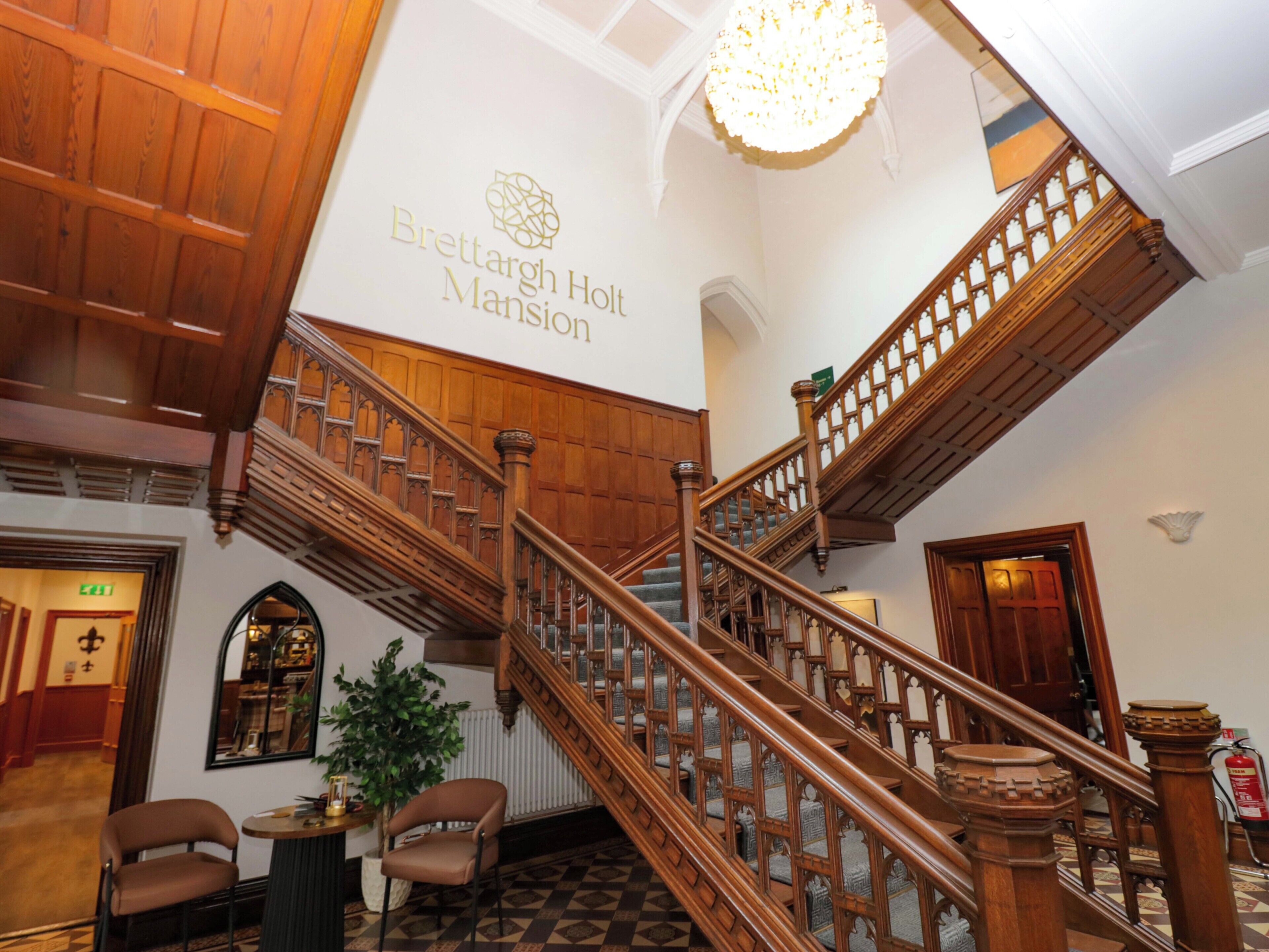 Una majestuosa escalera con barandillas de madera y un candelabro, que conduce a un techo alto con un letrero que dice 'Brettargh Holt Mansion'.