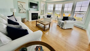 A living room with a fireplace, white sofas, a coffee table, and large windows with a view of the water. - Coastal Lines Retreat Ocean Views Sleeps 9 (Cocagne)