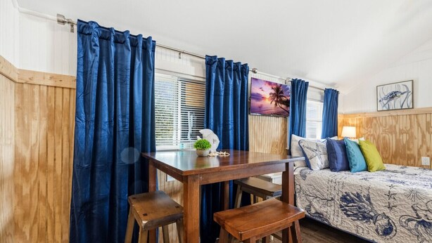 Ein Zimmer mit Holztisch, zwei Hocker, einem Bett mit bunten Kissen, einem Fenster mit Jalousien und einem Wandbild in einem Rahmen.