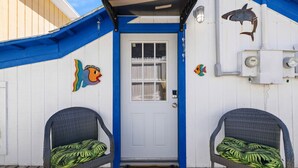 Eine weiße Tür mit blauem Rahmen, zwei Korbstühle mit grünen Kissen, ein Holzboden und Fisch-Wanddekoration.