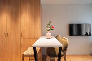 Un comedor con una mesa blanca, sillas de madera, un banco tejido, un florero con flores y un televisor de pantalla plana montado en la pared.
