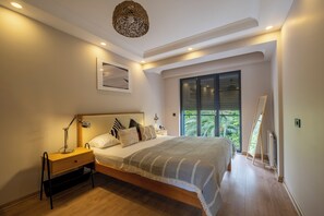 Ein Schlafzimmer mit Bett, Nachttisch, Lampe und Blick auf Grün hinter einem Fenster.