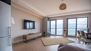A modern living room with a flat-screen TV, wooden flooring, a large window with a view of the sea, and a dining area with chairs. - Bosphorus View 2BR Luxury Apartment - Parking & Elevator (İstanbul)