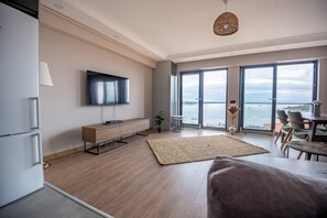 Ein modernes Wohnzimmer mit Flachbildfernseher, Holzboden, einem großen Fenster mit Meerblick und einem Essbereich mit Stühlen.