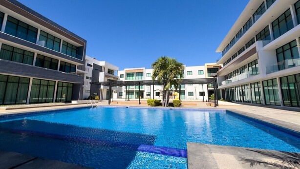 A modern building with a central swimming pool, surrounded by glass windows and a palm tree. - Golden Zone Escape: Stylish New Condo Steps from the Beach & Everything You Love (Mazatlan)