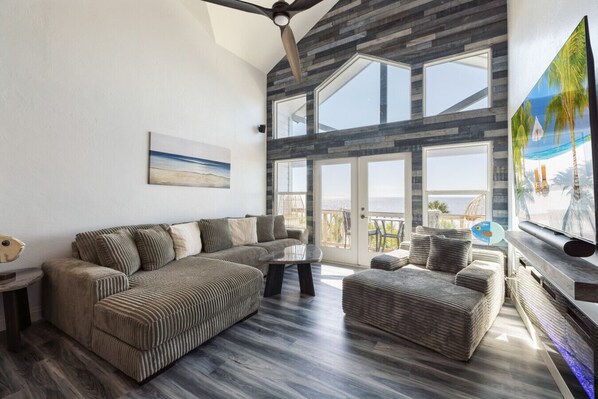 Ein modernes Wohnzimmer mit einem Ecksofa, einem Couchtisch und einem Fernseher. Große Fenster bieten Blick auf den Ozean.
