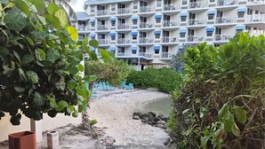 Et hotel ved stranden med en sti af sand, der fører ned til vandet, omgivet af grønt og blå stole.