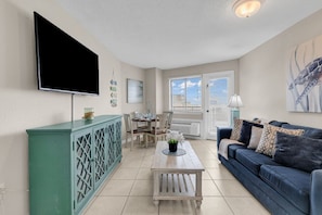 A living room with a flat-screen TV, a dining table, chairs, a sofa, and a window with blinds. - Bluewater Resort 1308 (Myrtle Beach)