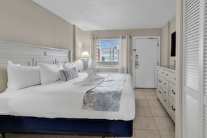 A neatly arranged hotel room with a large bed, a bedside table with a lamp, a wardrobe, and a door leading to another room. - Bluewater Resort 1308 (Myrtle Beach)