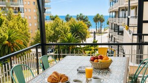 A balcony with a table set for breakfast, including croissants, fruit, and coffee, overlooking a beach and palm trees. - T3 6 pers - Sea view - Beach 1 min away - Terrace - WiFi fibre 1Gb - PortAventura (Salou)