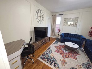 A living room with a TV, a blue sofa, a coffee table, a patterned rug, a clock, and a window with curtains. - Entire private 1 bedroom apartment in Brookline (Brookline)