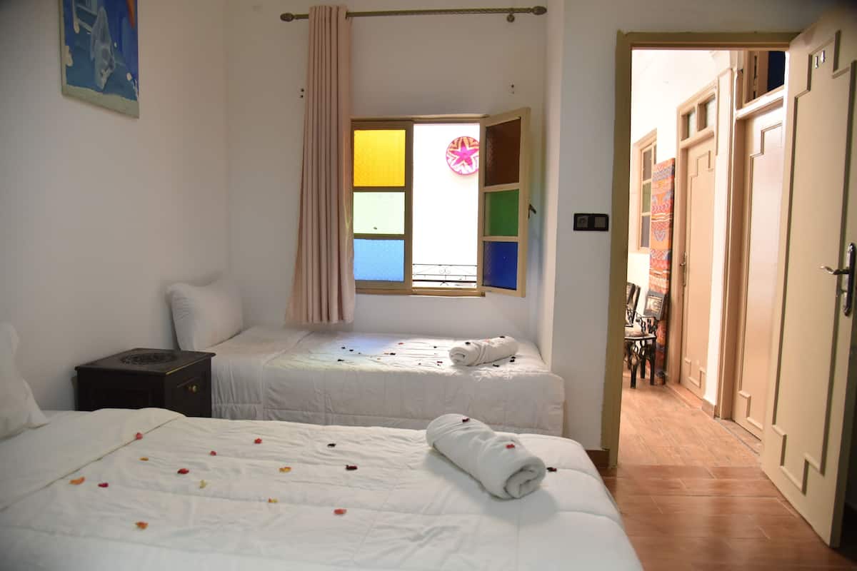 Ein Zimmer mit zwei Betten, einem Fenster mit farbigen Fensterscheiben, einem Vorhang und einer Tür, die in ein weiteres Zimmer führt.