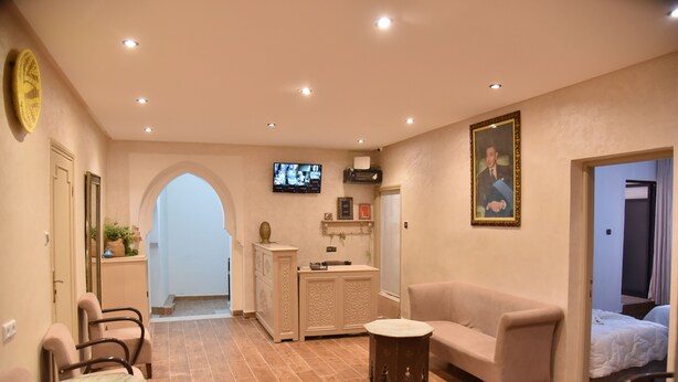 Quarto de hotel com sofá, mesa redonda pequena, televisão fixada na parede, um quadro e porta que dá para outro quarto.
