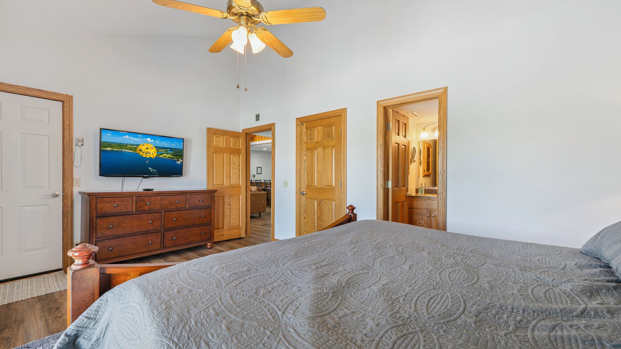 A bedroom with a bed, a wooden dresser, a television on the wall, and a ceiling fan.