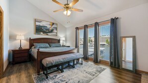 Un dormitorio con cama de madera, lámparas de noche, ventilador de techo, ventana grande con cortinas y vistas a la ciudad.