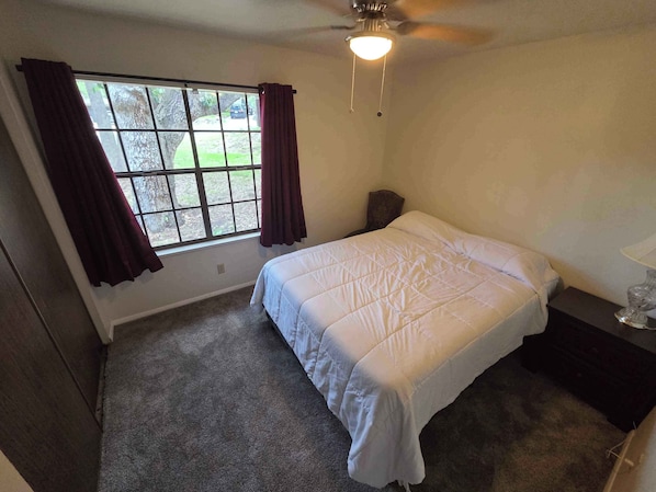 A bedroom with a bed, a window with curtains, a ceiling fan, and a nightstand with a lamp.