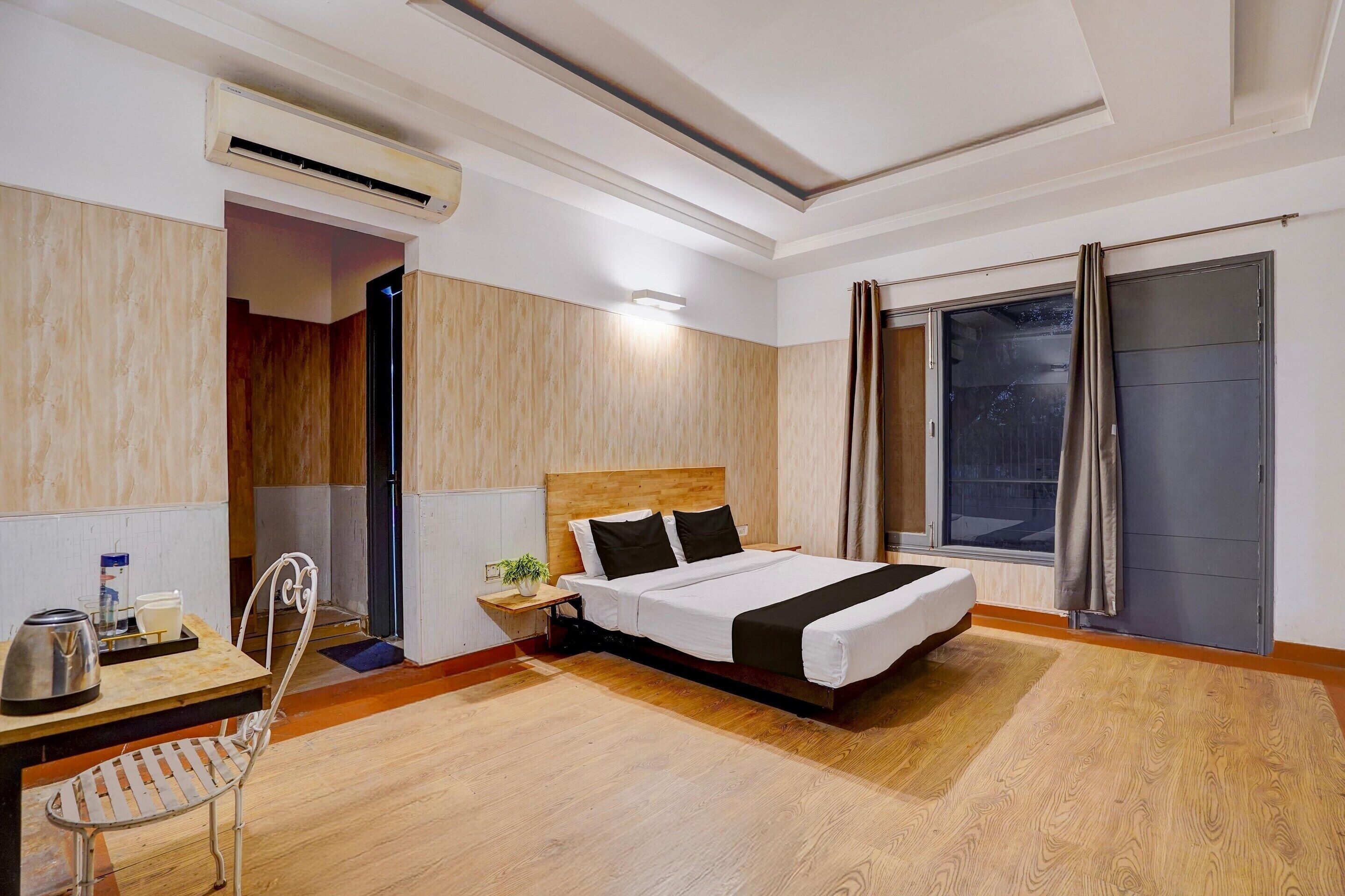 A hotel room with a bed, a desk with a chair, an air conditioning unit, a window with curtains, and a view of the outside.