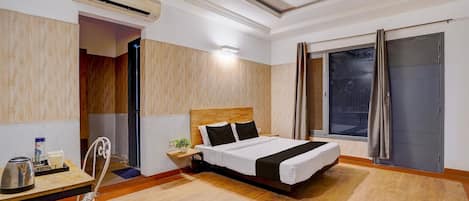 A hotel room with a bed, a desk with a chair, an air conditioning unit, a window with curtains, and a view of the outside.