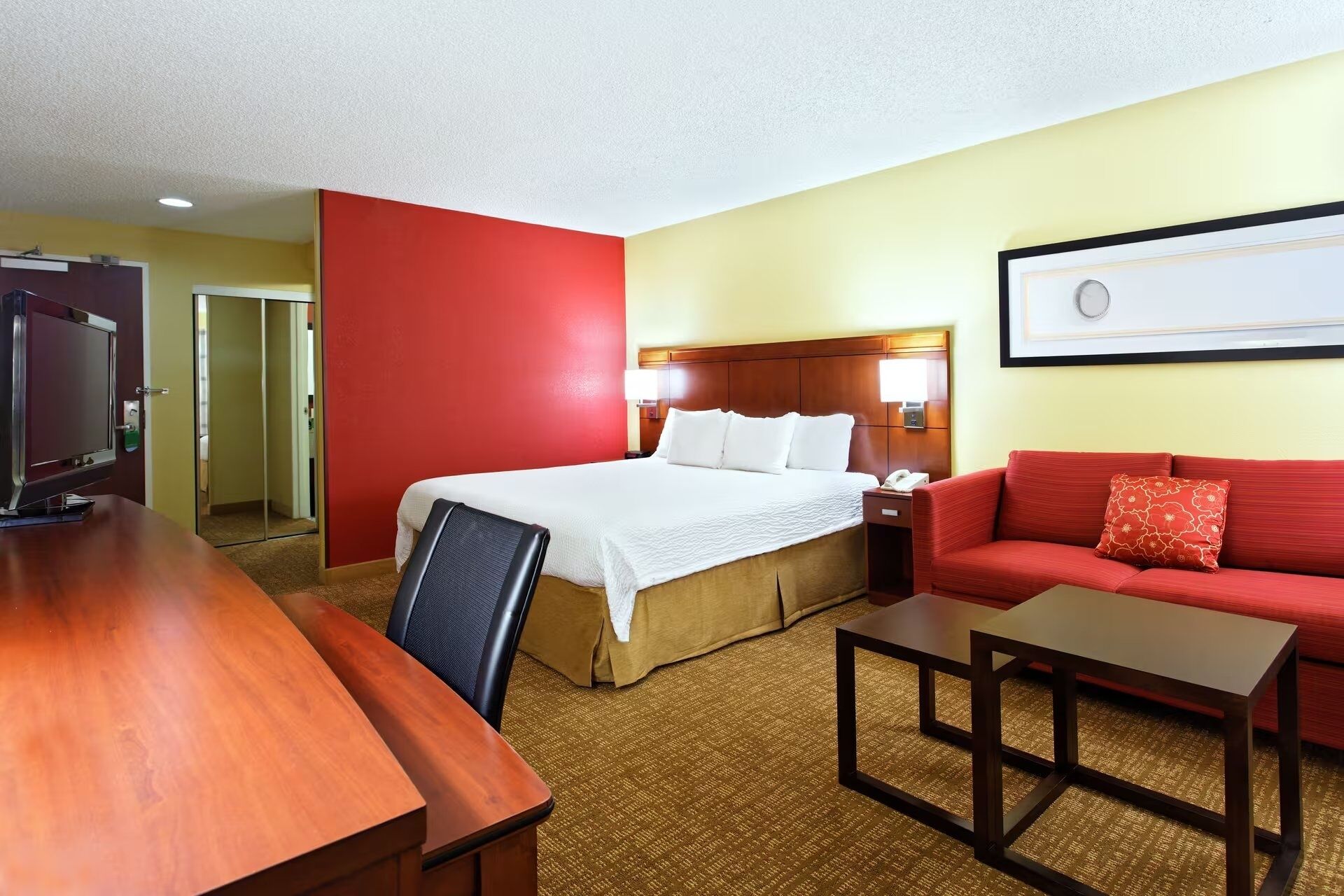Habitación de hotel con una cama, un sofá rojo, un escritorio de madera y una televisión.