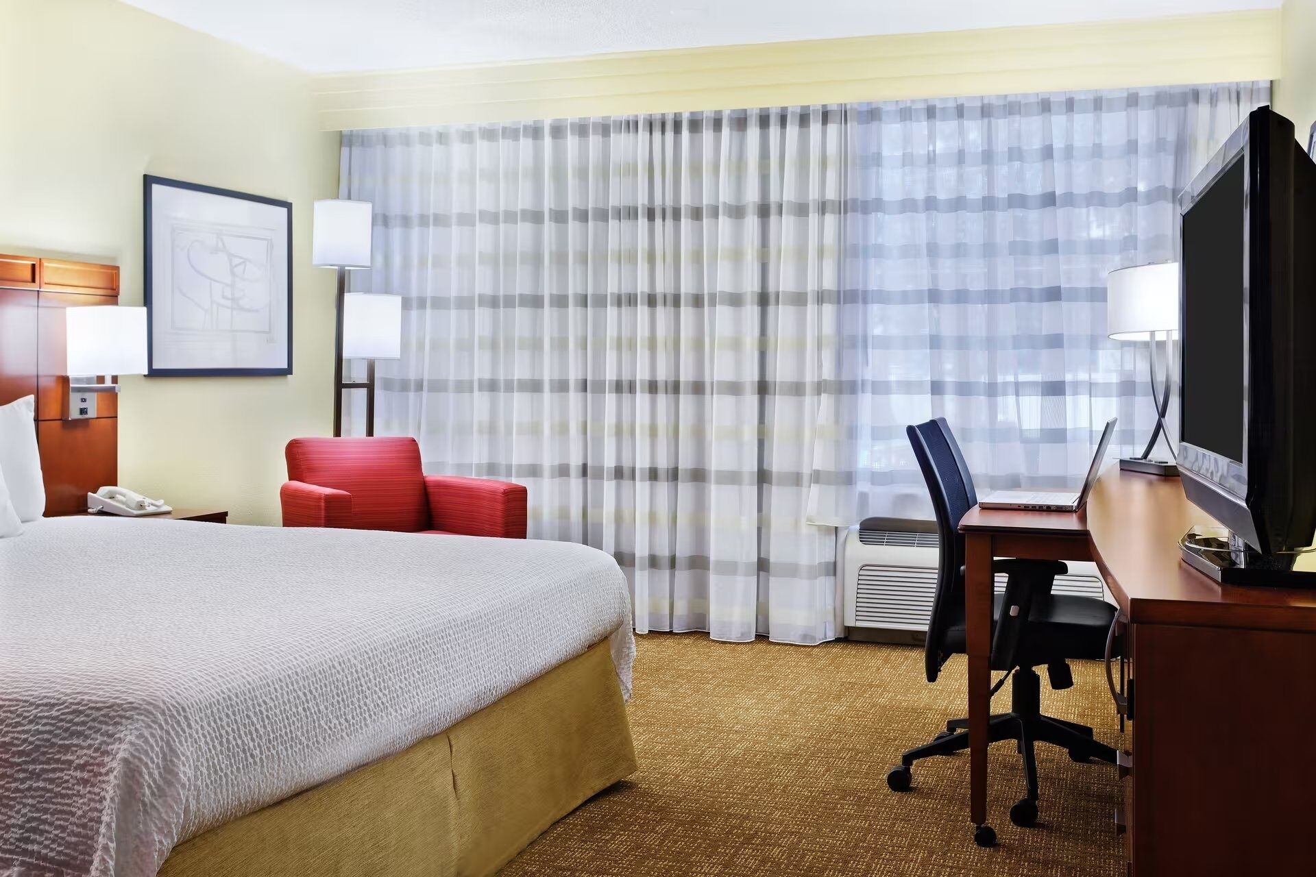 Habitación de hotel con una cama grande, un escritorio con una computadora portátil, una silla roja y un televisor.