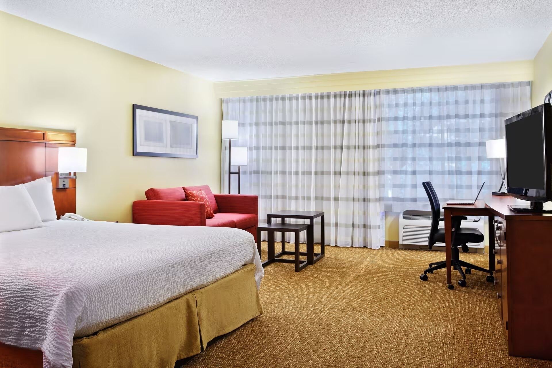Habitación de hotel con una cama grande, un sofá rojo, un escritorio con silla, televisor y ventana con cortinas.