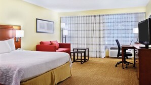 A hotel room with a large bed, a red sofa, a desk with a chair, a television, and a window with curtains. - Modern Houston Corner Room with King Bed, Sofabed & Pool Access (Houston)