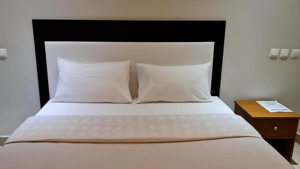 A neatly made bed with a white headboard and pillows, a wooden nightstand with a lamp, and a small table with a menu or information card.